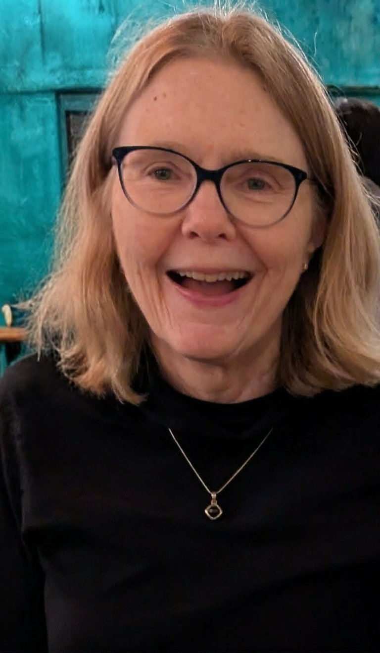 Woman with shoulder-length reddish-brown hair and glasses smiling at camera, wearing black shirt with pendant necklace